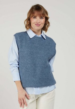 Jeune femme aux cheveux courts et ondulés portant un gilet sans manches en laine bleu par-dessus une chemise à rayures bleu clair à boutons et un pantalon beige.