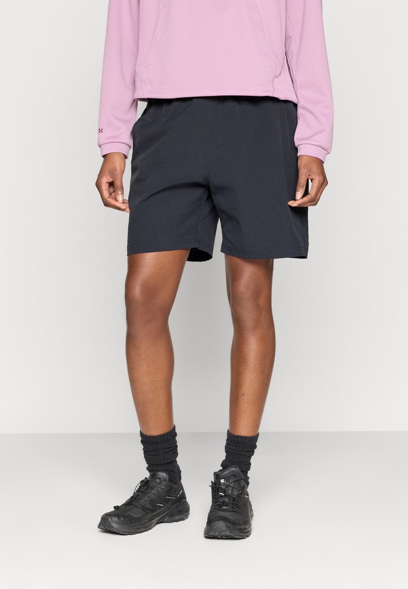 Μαύρο αθλητικό σορτς με λεία υφή, μήκος πάνω από το γόνατο, συνδυασμένο με μαύρα παπούτσια και υπερθετικό ανοιχτό μωβ μακρυμάνικο τοπ.