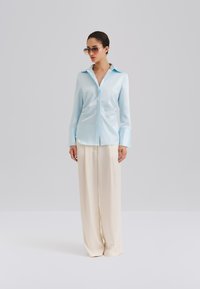 Ανοιχτό μπλε σατέν πουκάμισο με μακριά μανίκια, αιχμηρό γιακά, κουμπιά μπροστά και άνετη εφαρμογή, σε συνδυασμό με πλατύχωρα μπεζ παντελόνια.