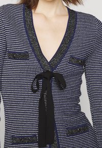 Gros plan d’un cardigan pied-de-poule bleu et blanc avec bordure noire, poches à rabat boutonnées et un nœud noir noué sur la poitrine.