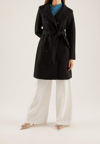 Černý dvouřadý kabát s širokým límcem a páskem, vyrobený z měkkého materiálu. V kombinaci s bílými kalhotami s širokými nohavicemi a zdobenými botami.