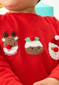 Pull en tricot rouge avec trois motifs festifs brodés : un renne avec des bois, un pudding de Noël et le Père Noël, chacun avec des accents colorés.