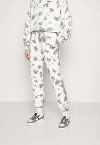 Person som bär svettbyxor och sweatshirt i vitt och grått batikmönster med svarta Nike-logotyper, i kombination med vita strumpor och vita, röda och grå Nike-sneakers.