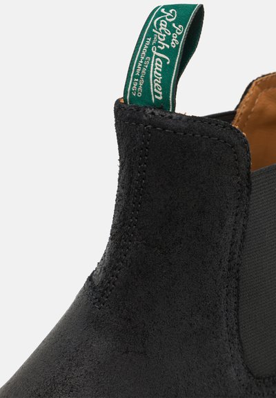 Botín de ante negro con paneles laterales elásticos y una lengüeta de tela verde con logo blanco. Forro interior suave, diseño elegante.