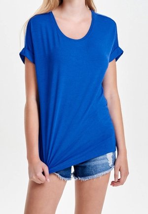 Mujer con una camiseta azul lisa de manga corta y shorts de mezclilla desgastados, sujetando el dobladillo de la camiseta con una mano.