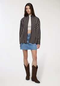 Camicia oversize a scacchi bianco e nero, top corto bianco, gonna di jeans azzurri chiari, stivali da cowboy marroni con design in rilievo, su sfondo bianco.