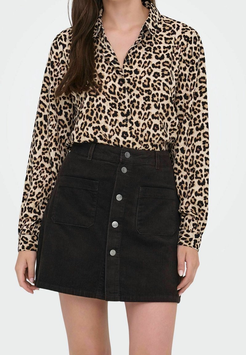 Lange mouwen blouse met luipaardprint, gecombineerd met een hoog opgesneden zwarte ribfluwelen rok met voorzakken en metalen knoopdetails.