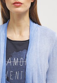 Světle modrý kardigan s otevřeným předním dílem, vyrobený z tenké, měkké látky, přehozený přes tmavé tričko s bílým textem a texturovaným vzorem.
