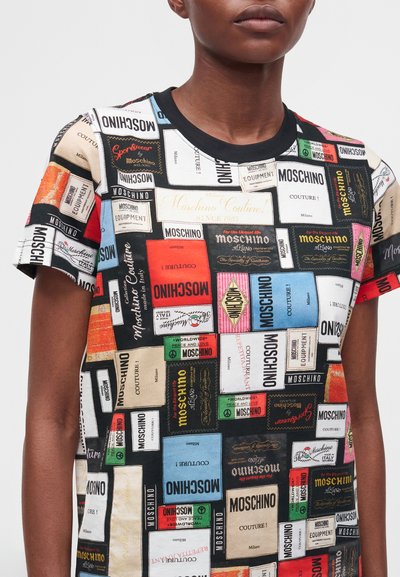 Personne portant un t-shirt patchwork multicolore avec le logo Moschino et différentes étiquettes de marque en rouge, bleu, noir, blanc et beige.