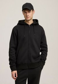 Czarna bluza z kapturem i zamkiem błyskawicznym, z przednią kieszenią, kapturem ze sznurkiem i ściągaczami na rękawach. Nosi ją w komplecie z pasującą czarną czapką. Gładka tekstura materiału.
