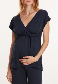 Marineblause mit kurzen Ärmeln für Schwangere, einer gebundenen Taille und weichem Stoff, kombiniert mit passenden marineblauen Unterteilen. Bequeme und funktionale Gestaltung.