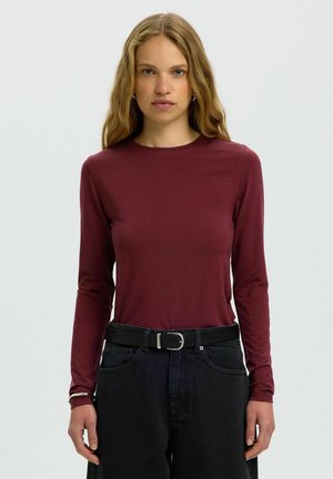 Femme aux longs cheveux blonds ondulés portant un haut cintré bordeaux à manches longues et un jean noir taille haute avec une ceinture noire, debout devant un fond uni.