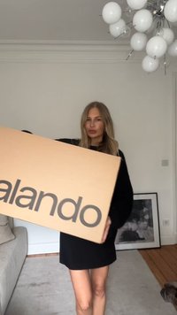 Rjavo kartonsko škatlo z napisom "zalando" v velikih sivih črkah, držano pokončno, v prostoru s svetlimi stenami in lesenim tlakom.