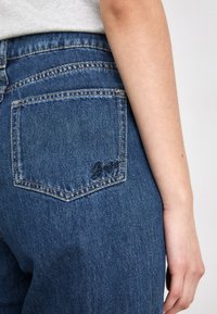 Donkerblauwe denim jeans met een klassieke snit, voorzien van een achterzak geborduurd met een cursief logo en zichtbare stikdetails.