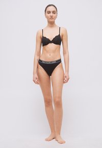 Γυναίκα όρθια, ξυπόλητη, φοράει μαύρο σουτιέν δαντέλας και μαύρα εσώρουχα με το λογότυπο Polo Ralph Lauren, μπροστά σε απλό φωτεινό φόντο.