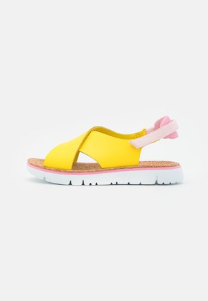 Gul sandal med åben tå og krydsede remme, korksål, pink ankelrem og hvid fleksibel sål.