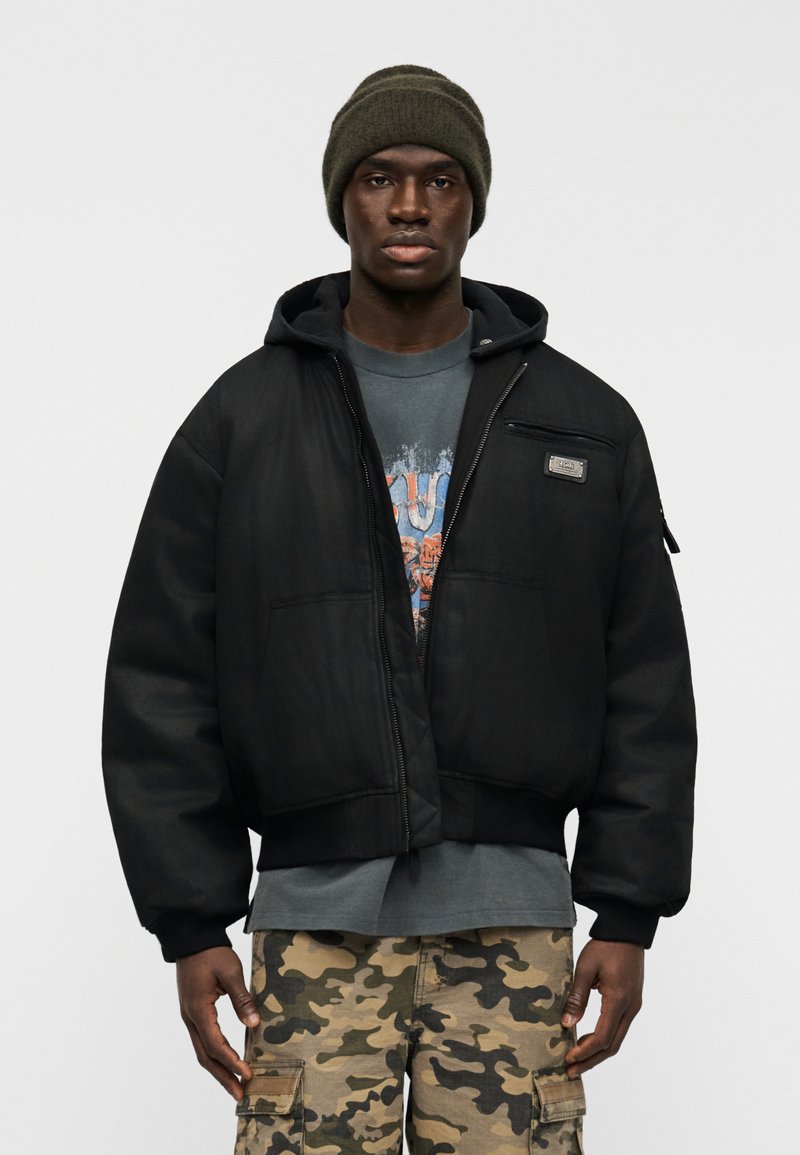 Μαύρο μπουφάν μπόμερ με φερμουάρ σ το μπροστινό μέρος, ριμπ μανσέτες και κουκούλα, φορεμένο πάνω από ένα γραφικό t-shirt και κάπρι παντελόνια καμουφλάζ. Χακί σκούφος στην κορυφή.