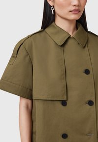 Oliwkowy, wodoodporny trencz z krótkimi rękawami, dużym kołnierzem, podwójnym zapięciem z przodu oraz czarnymi guzikami. Posiada naramienniki.