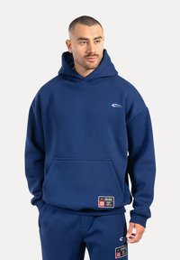 Mann in passendem blauen Kapuzenpullover und Jogginghose mit weißen und roten Logo-Patches, der leicht nach rechts schaut, vor einem schlichten Hintergrund.