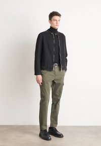 Chaqueta negra con cremallera sobre un cuello de tortuga negro, pantalones cargo oliva y zapatos negros. Diseño minimalista con una textura suave y un corte ajustado.