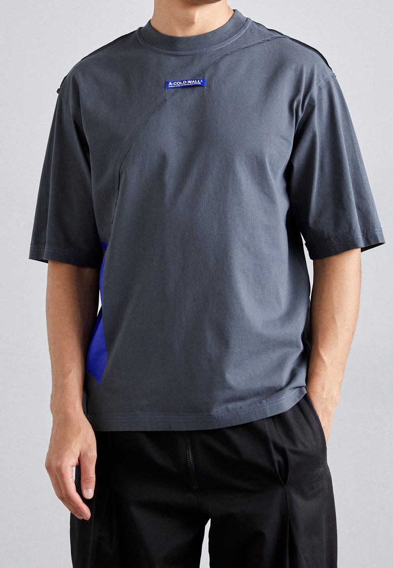 Homme portant un t-shirt gris foncé à manches courtes avec un petit écusson logo bleu "A-COLD-WALL*" sur la poitrine et un pantalon noir.