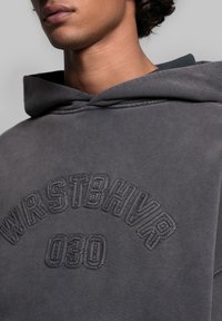 Sweat à capuche gris avec un grand logo brodé, affichant le texte « WRSTBHV » et « 030 » dans un point surélevé et texturé. Tissu doux et décontracté.