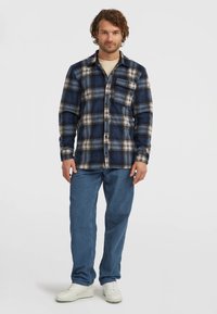 Flanellen overhemd in marineblauw en crèmekleurig ruitpatroon, met knoopsluiting aan de voorkant, één borstzak. Lichtblauwe denim broek en witte sneakers.