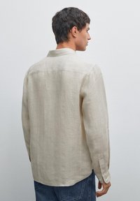 Man facing sideways wearing a long-sleeve beige linen shirt and blue jeans against a plain light gray background.