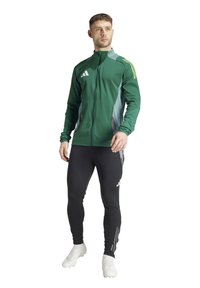 Giacca verde con zip e accenti grigi, tre righe bianche sulle maniche, abbinata a leggings neri con logo e dettagli a righe.