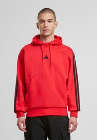 Röd huvtröja med framficka, svart Adidas-logotyp och svarta ränder på ärmarna. Tillverkad av mjukt tyg med snodd i huvan.