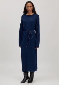 Marineblaues langärmliges Kleid mit lockerem Schnitt, tailliert mit einem Bindegürtel und weicher Textur. Getragen mit knielangen schwarzen spitzen Stiefeln.