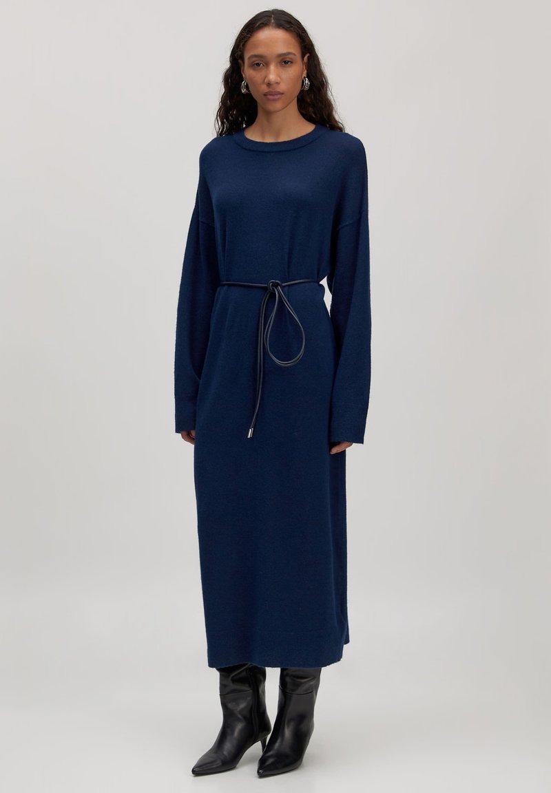 Marineblaues langärmliges Kleid mit lockerem Schnitt, tailliert mit einem Bindegürtel und weicher Textur. Getragen mit knielangen schwarzen spitzen Stiefeln.