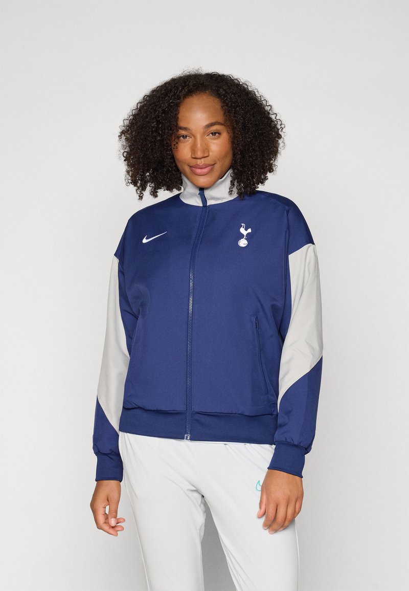 Marinblå zip-up jacka med grå detaljer och en vit Tottenham-logotyp på bröstet; har en hög krage och sidofickor.