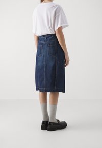 Žena v bílé tričko zasunutém do tmavě modré denimové midi sukně, šedých žebrovaných ponožkách a černých mokasínech se zlatými detaily, stojící bokem.