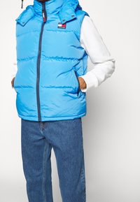 Sininen toppaliivi, jossa korkea kaulus, etuvetoketju ja sivutaskut. Rinnassa Tommy Jeans -logolla varustettu merkintätarra. Karkea, eristetty kangas.