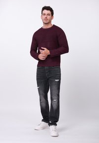 Pull en maille bordeaux avec des rayures texturées, associé à un jean noir déchiré et des baskets blanches. Le mannequin pose devant un fond uni.