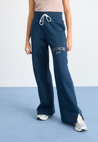 Kvinna i marinblå Nike sweatpants med vit snörning och sneakers, stående på blått golv mot vit vägg.