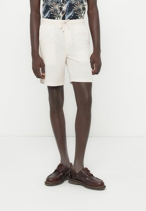 Pantalones cortos de lino en beige claro con cintura elástica y cordón. Combinados con una camisa negra estampada y zapatos de cuero marrón.