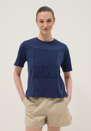 Jeune femme aux cheveux attachés portant un t-shirt bleu marine à manches courtes avec un motif carré brodé et un short beige, mains dans les poches.