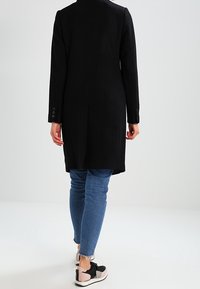 Musta pitkä takki, jossa on istuva siluetti, yksinkertainen nappisulku, pitkät hihat ja sileä pinta. Yhdistettynä sinisiin slim fit -farkkuihin ja mustiin lenkkareihin.