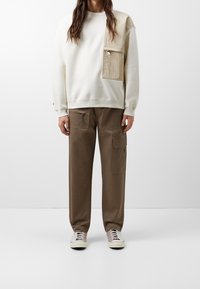 Sudadera blanca con un bolsillo de parche beige, combinada con pantalones cargo marrones y zapatillas grises. Presenta un diseño sencillo y texturas suaves.