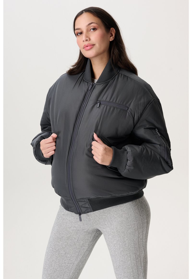 Eine Frau trägt eine dunkelgraue wattierte Bomberjacke mit Reißverschlusstaschen und hellgrauen, gerippten Leggings und steht vor einem weißen Hintergrund.