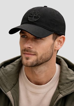 Uomo con leggera barba indossa un berretto nero con logo ad albero, giacca verde e maglia beige con collo rotondo, guarda leggermente verso destra su sfondo semplice.