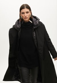 Černý péřový kabát s kapucí, vybavený zipovým zapínáním a prošívanou podšívkou, nošený přes černý rolák a kožené kalhoty.