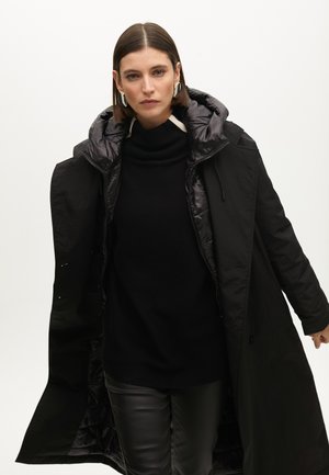 Abrigo de plumas negro con capucha, con cierre de cremallera y forro acolchado, llevado sobre un jersey negro de cuello alto y pantalones de cuero.