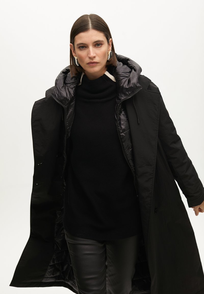Černý péřový kabát s kapucí, vybavený zipovým zapínáním a prošívanou podšívkou, nošený přes černý rolák a kožené kalhoty.