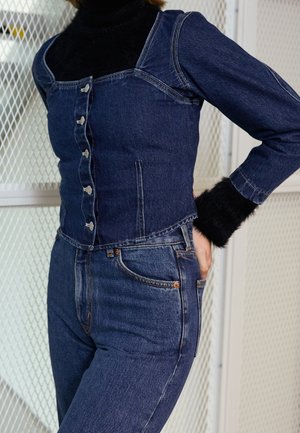 Personne portant une veste en denim foncé boutonnée sur un col roulé noir duveteux, associée à un jean en denim foncé assorti, debout avec une main sur la hanche.
