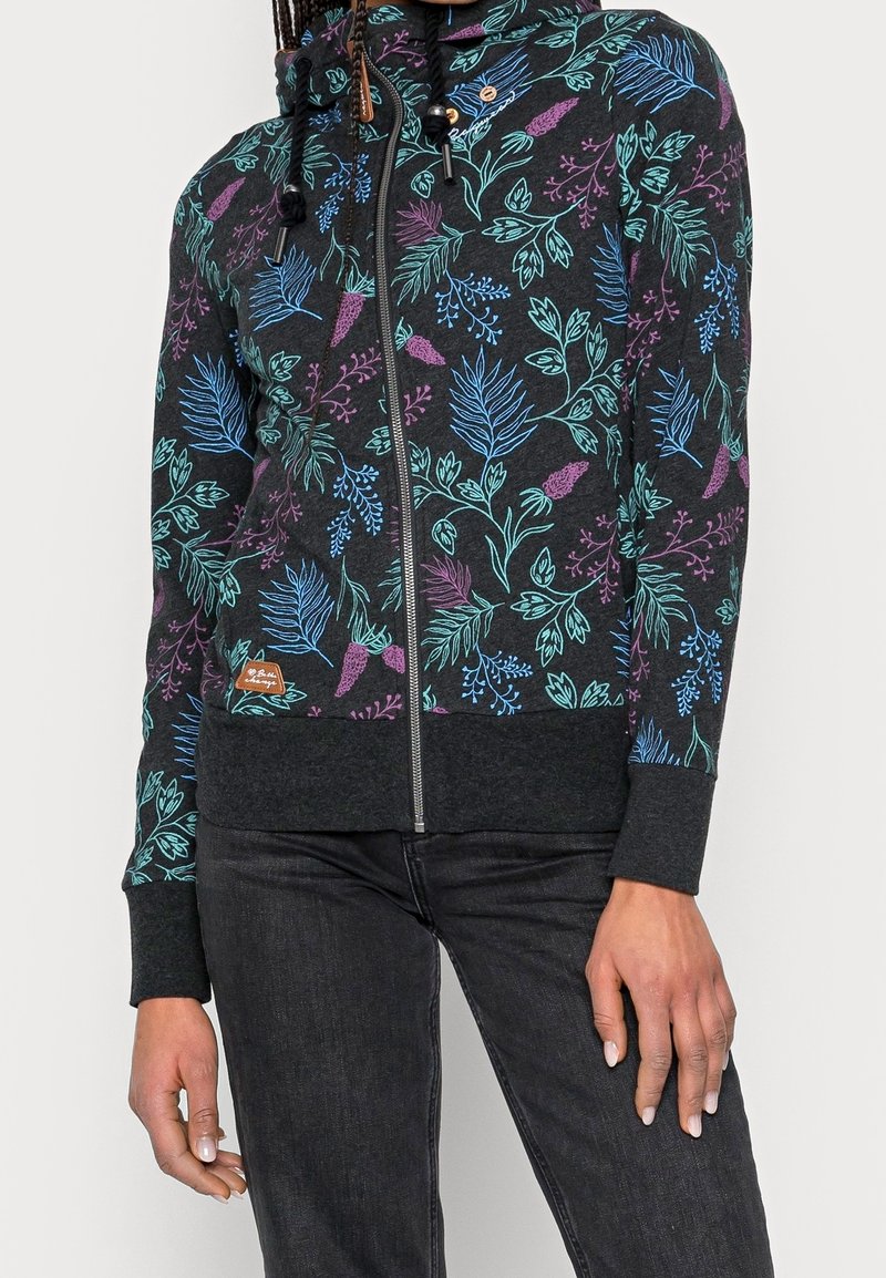 Femme portant un sweat à capuche noir zippé avec des motifs floraux bleus, verts et violets et un pantalon noir sur un fond uni.
