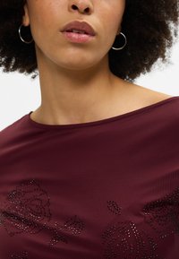 Burgundijas krāsu tops ar plašu dekoltē, kas rotāts ar ziedu motīviem, apdarināts ar melniem un sarkaniem rhinestones, gluda tekstūra un mīksta auduma.