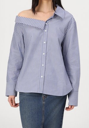 Mujer con camisa de rayas verticales azul y blanca, sin hombros, abotonada, combinada con vaqueros azules.